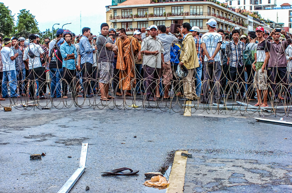 A large crowd of people standing behind a barbed wire barricade on a street, with some individuals appearing to be engaging in a discussion or protest.