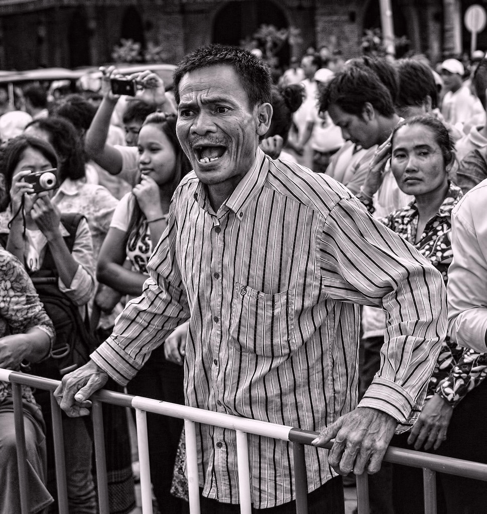 A crowd of people gathered at an event, with a man in the foreground shouting passionately. He is wearing a striped shirt and appears to be leaning on a barrier, while others around him watch intently.