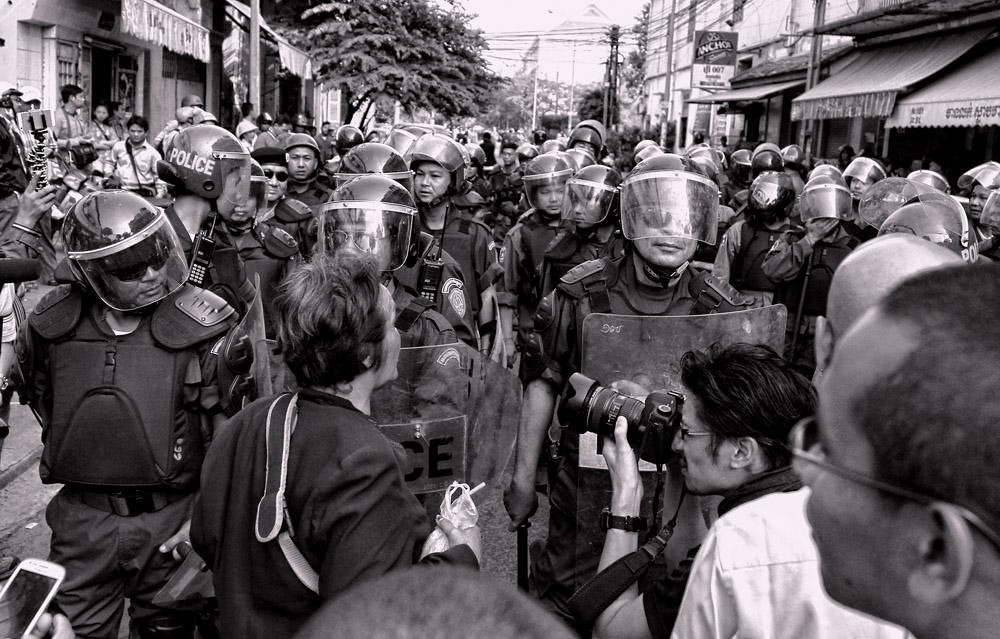 A black and white image of a tense standoff between police in riot gear and a woman speaking to them, with a crowd of onlookers and media present.