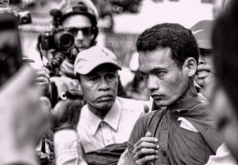 A tense moment captured in black and white, showing a young man surrounded by a crowd and cameras, appearing contemplative and serious.