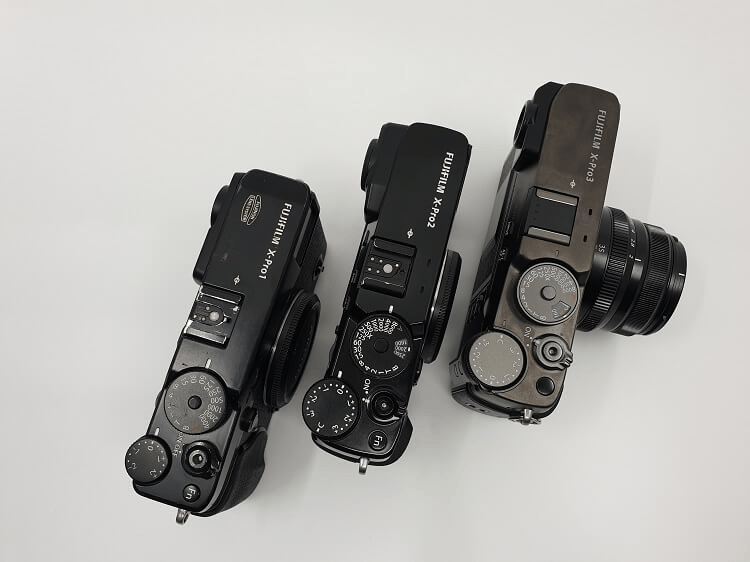 Three Fujifilm X-series cameras lined up, showcasing their top views. The models include the X-Pro1, X-Pro2, and X-Pro3, all in black with various dials and settings visible.