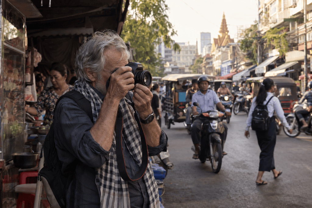 A man with gray hair and a scarf around his neck is photographing a bustling street scene with a camera, surrounded by street vendors and traffic in an urban setting.