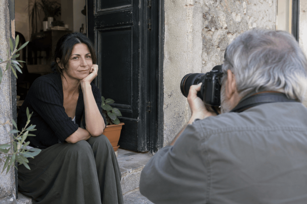 A woman sitting on a doorstep with a thoughtful expression, while an older man photographs her using a camera.