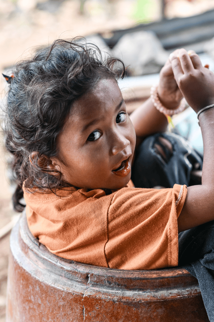 A young child with curly hair wearing an orange shirt, sitting in a large pot, looking towards the camera with a playful expression.