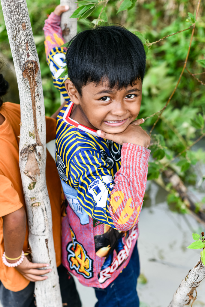 A smiling young boy with short black hair, wearing a colorful patterned shirt, poses playfully by a tree near water.