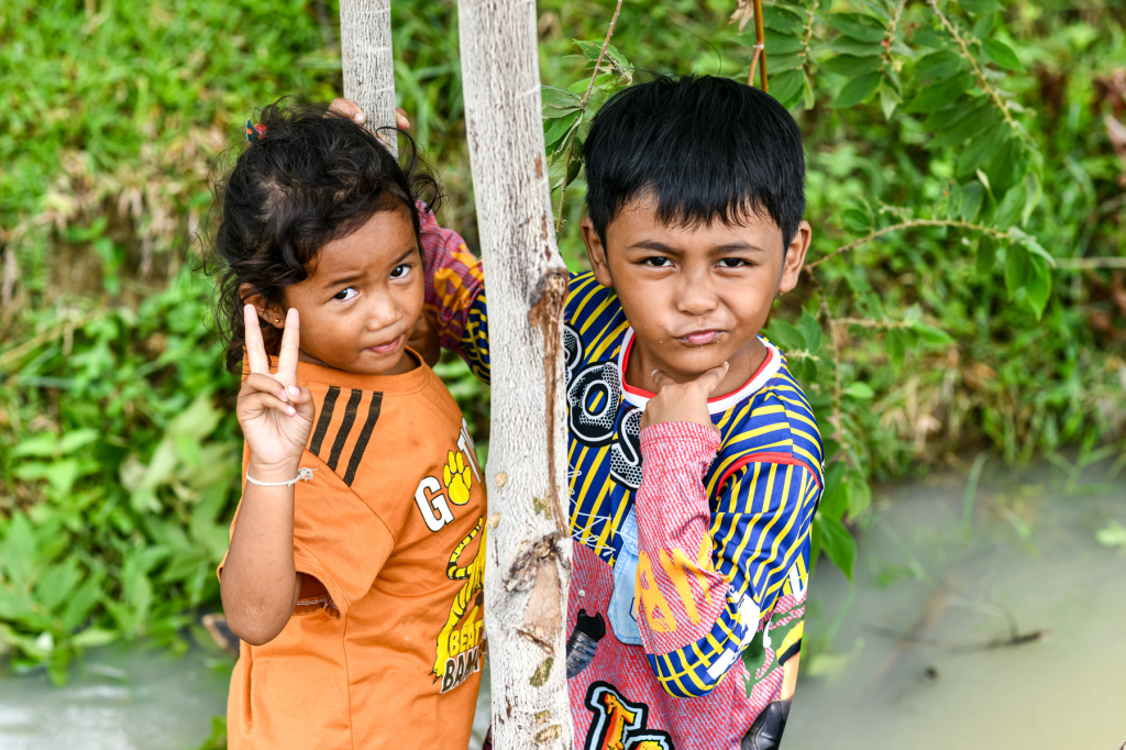 Two children posing playfully outdoors, with one girl making a peace sign and a boy pouting, surrounded by greenery.
