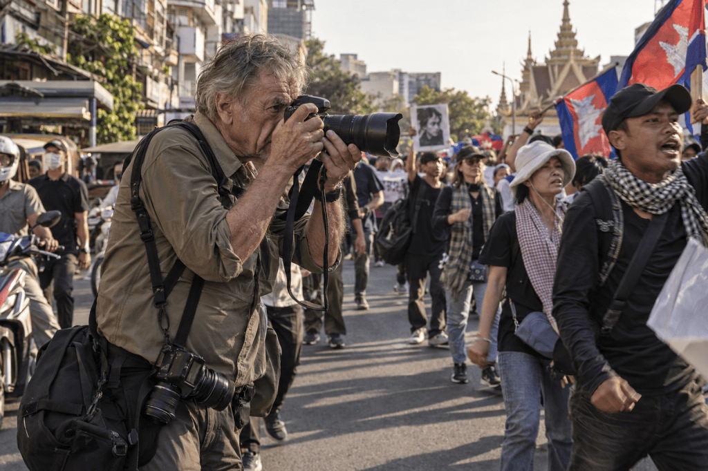 A photographer capturing images at a protest with a large crowd of demonstrators holding flags and banners in a city street.