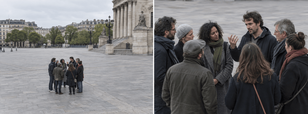 A group of people engaged in a conversation in a public square, with a building visible in the background and cloudy skies above.