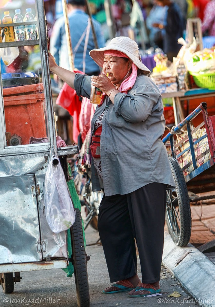 An elderly woman stands beside a food cart, sipping a drink. She wears a wide-brimmed hat and a gray shirt, with a scarf around her neck. The bustling market scene in the background features colorful stalls and people.