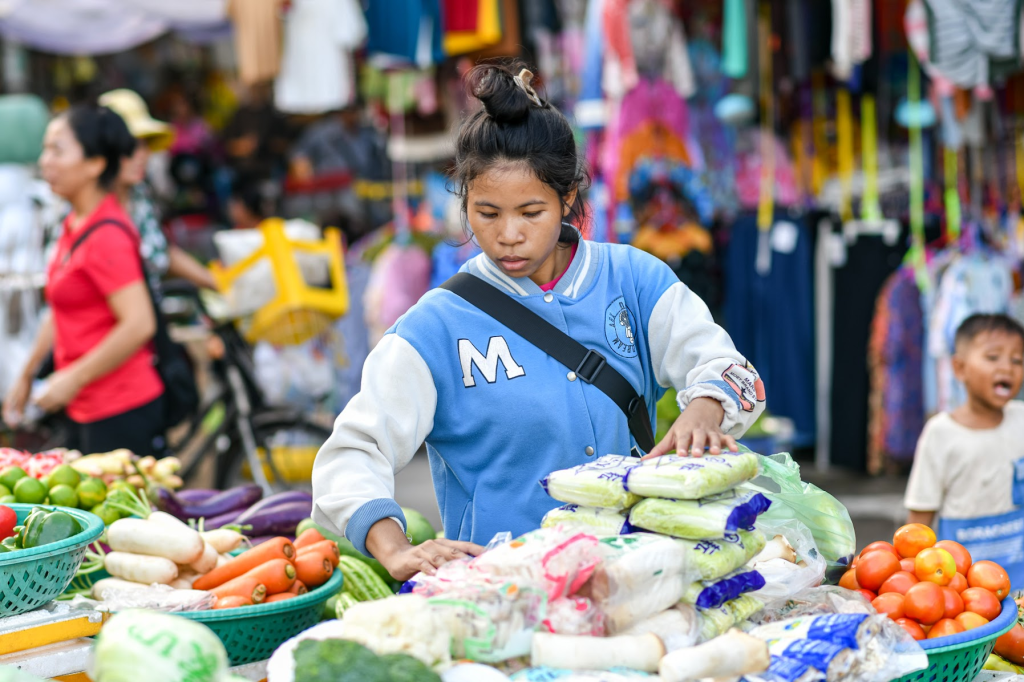 A young girl in a blue jacket arranging fresh vegetables and fruits at a market stall, with shoppers and colorful merchandise in the background.