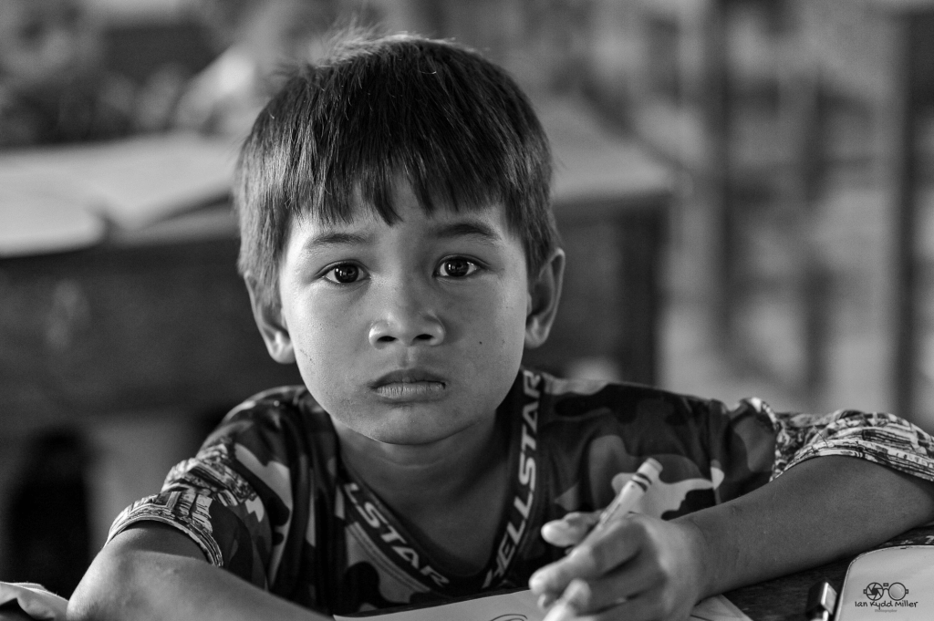 A close-up of a young boy with a serious expression, sitting at a desk, holding a pen, in a classroom setting.
