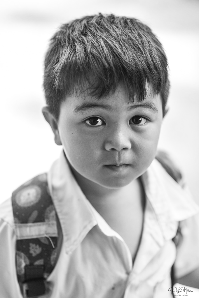 A young boy with short hair and a serious expression is looking directly at the camera. He is wearing a white shirt and has a backpack with a colorful strap.