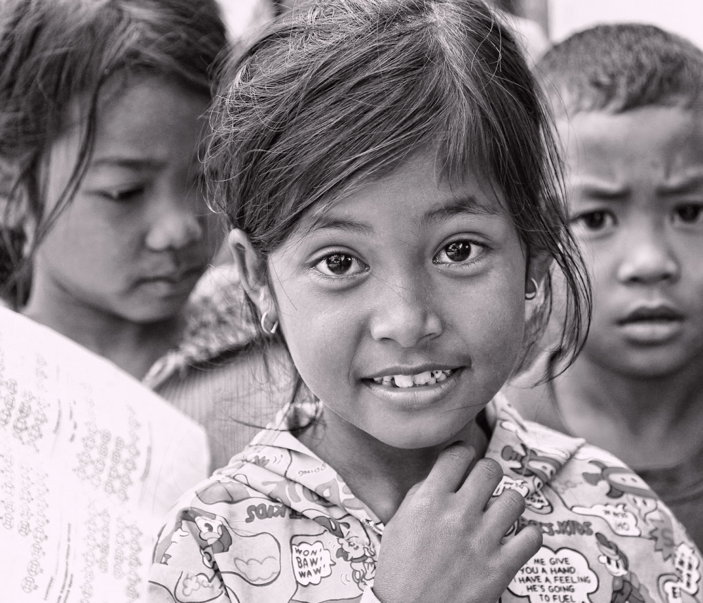A smiling young girl in a colorful shirt, with dark hair and striking eyes, stands in the foreground, while two other children appear slightly blurred in the background.