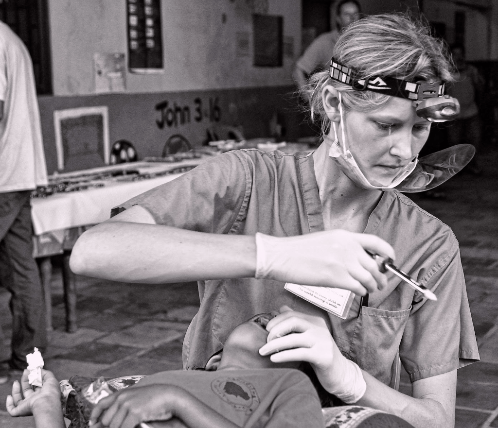 A healthcare worker in surgical scrubs and gloves, using a medical tool to attend to a child in a clinical setting.