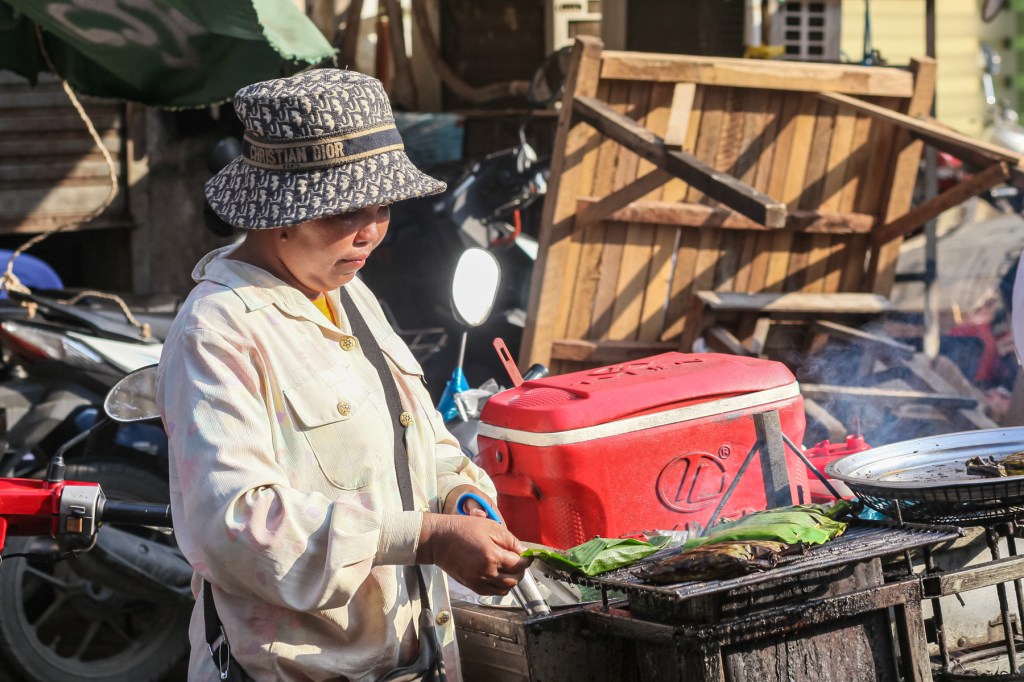 A woman wearing a patterned hat and a light-colored shirt prepares food at a street vendor stall, with smoke rising from a grill and a cooler nearby.