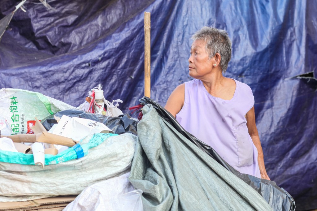 An elderly woman wearing a lilac shirt rummaging through a pile of trash bags and recyclable materials under a blue tarp.