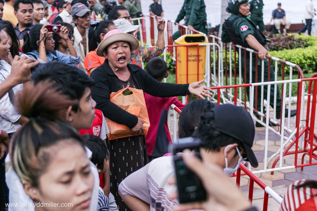 A crowd of people gathered, with a woman in the foreground passionately speaking or shouting, holding a bag. Several others are taking photos and observing the scene, while security personnel are visible in the background.
