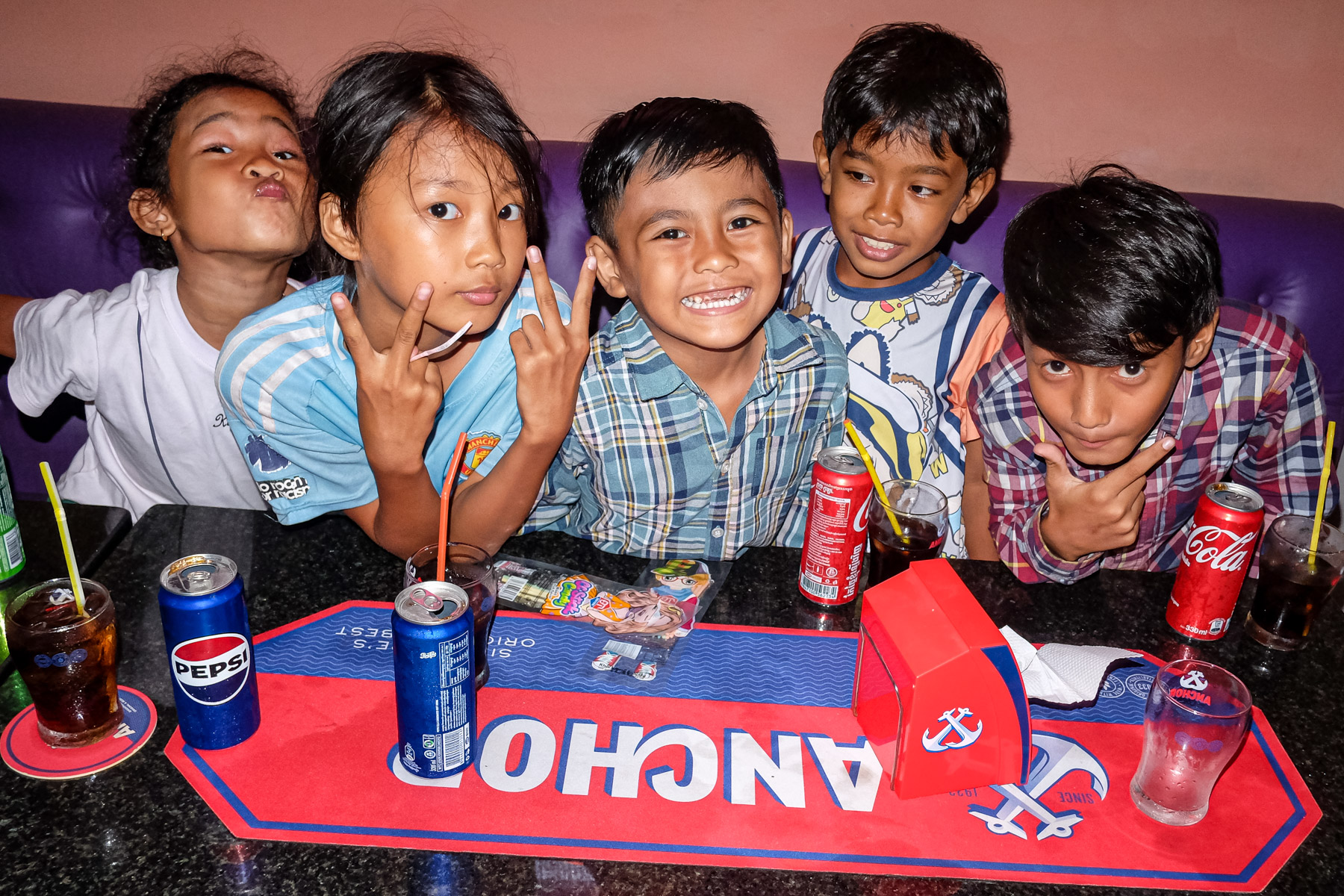 A group of five children sitting at a table with drinks, smiling and posing playfully for the camera in a colorful setting.