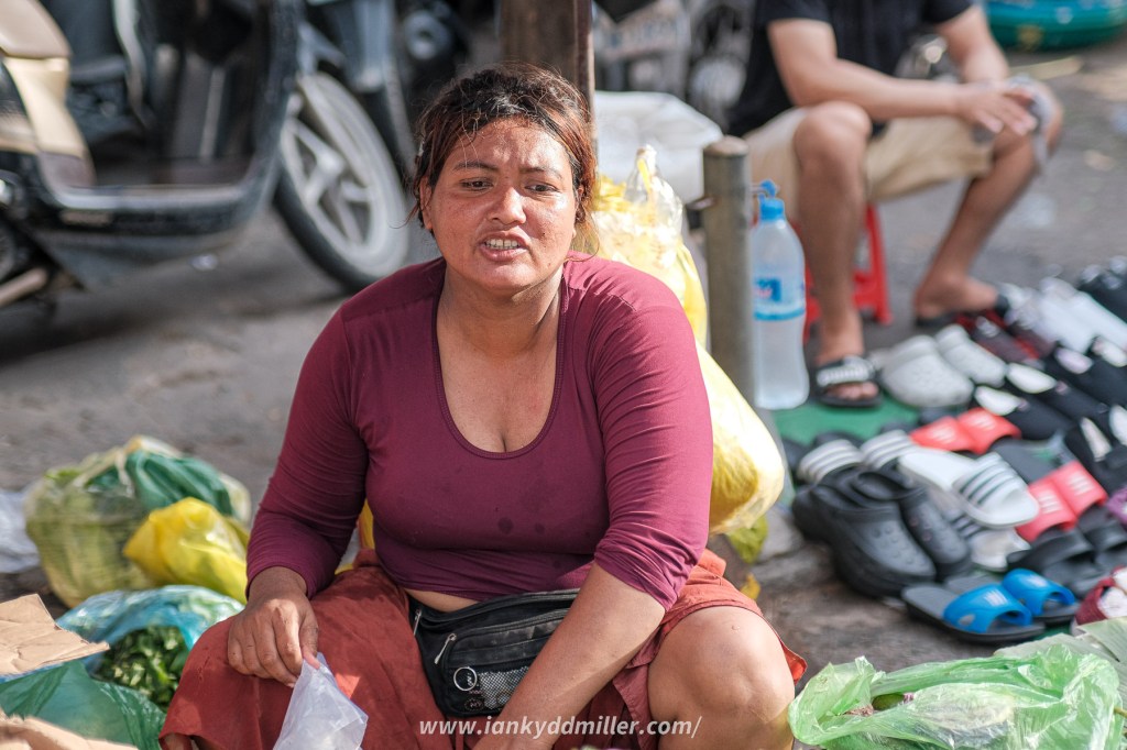 A woman sitting on the ground surrounded by bags of produce, looking into the camera with a serious expression. In the background, there are other people and various items for sale, including footwear.