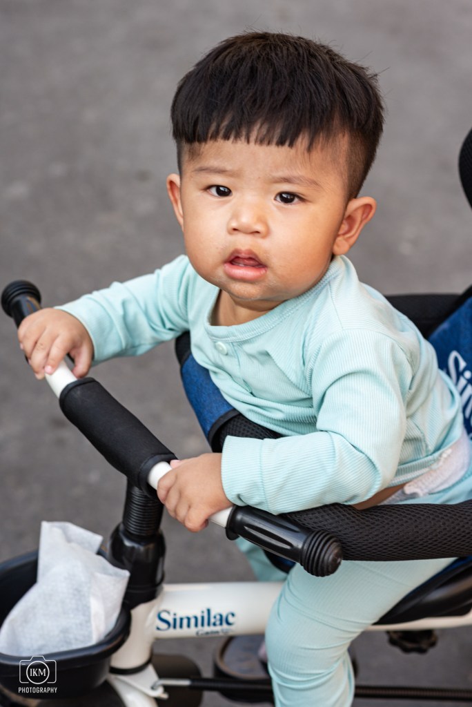 A young child wearing a light blue outfit, sitting on a tricycle and holding the handlebars, looking towards the camera.