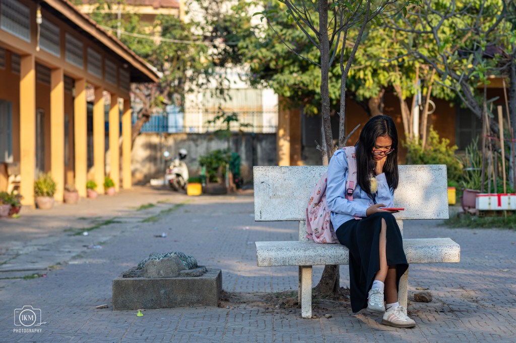 A young girl sitting on a stone bench in an outdoor area, looking at her phone. She wears a blue shirt, black skirt, and has a pink backpack. Surrounding her are trees and buildings.