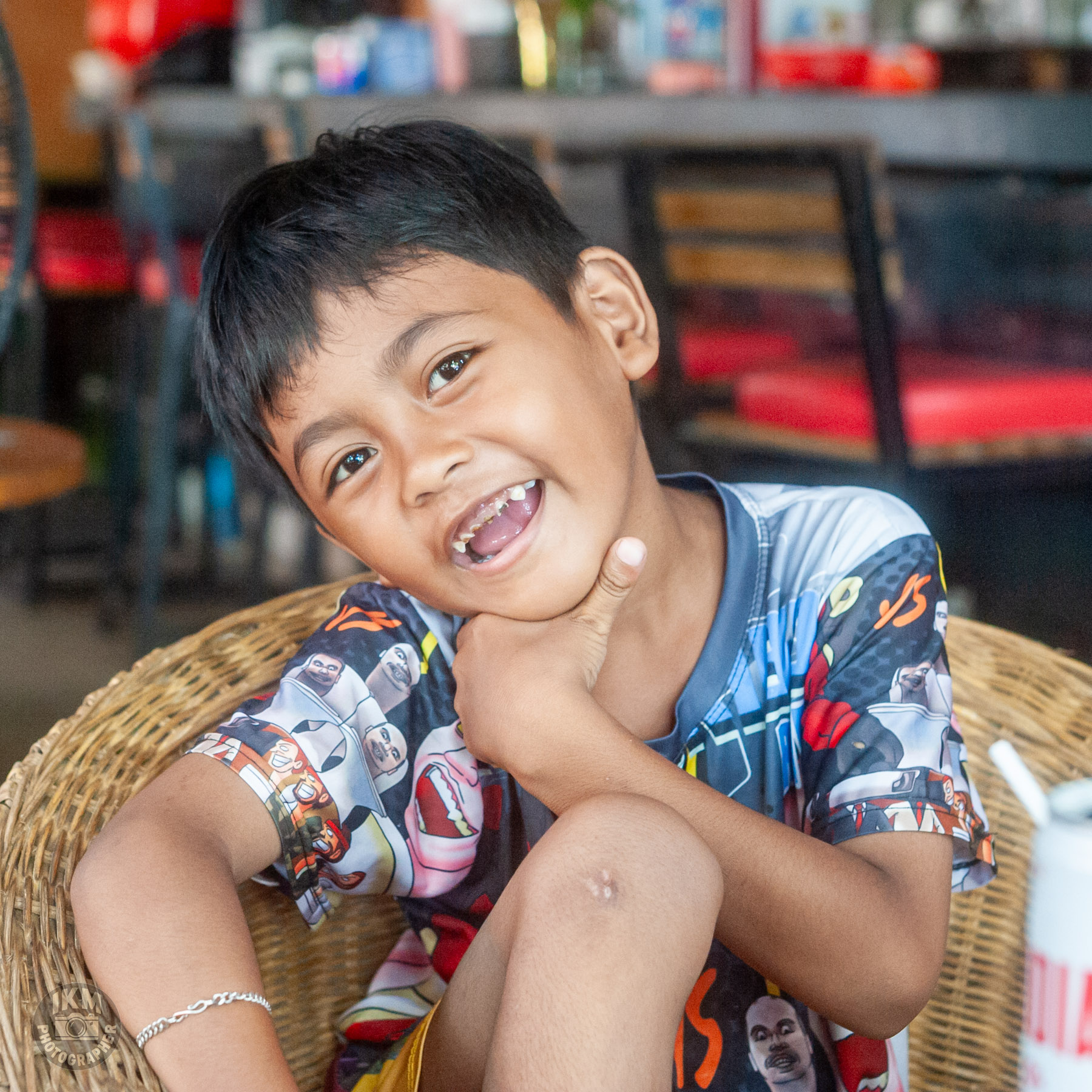 A smiling young boy with short black hair, sitting casually in a wicker chair, wearing a colorful graphic t-shirt, and playfully resting his chin on his hand.