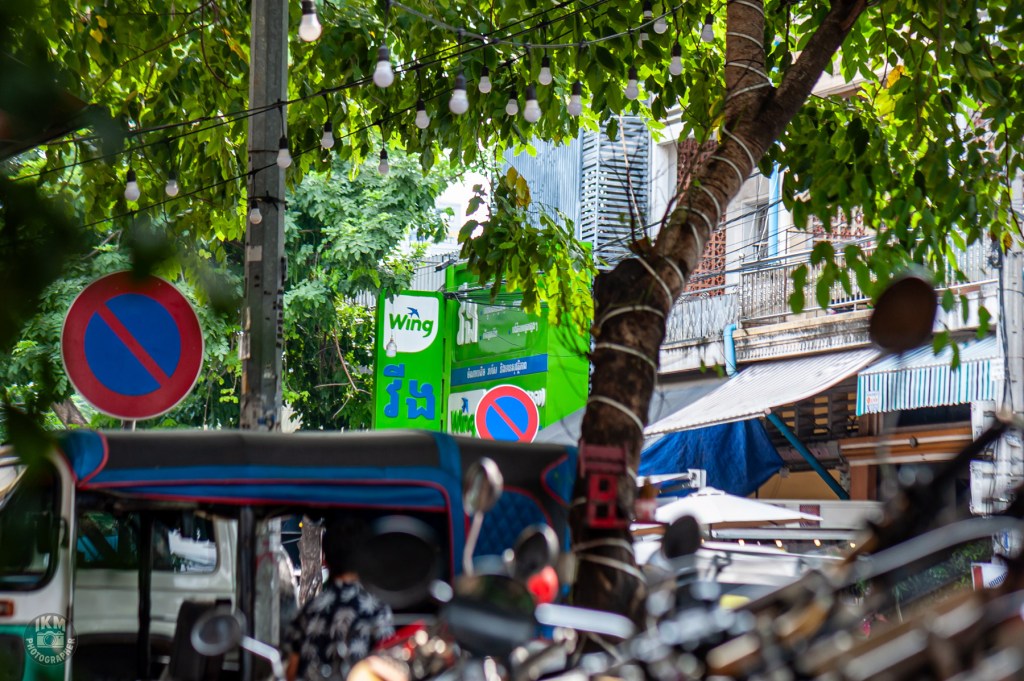 A bustling street scene featuring a tree with vibrant green leaves, a 'no parking' sign, and a prominent advertisement for Wing. The background includes buildings and various parked vehicles.