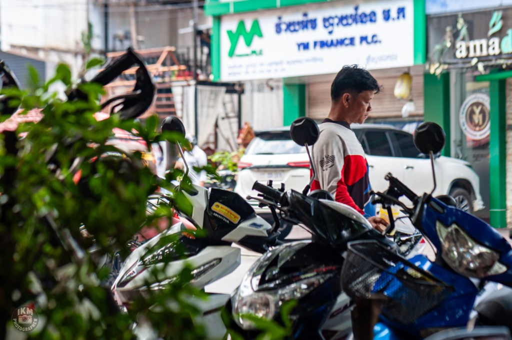 A young man wearing a red and white long-sleeve shirt is standing near a row of parked motorcycles, surrounded by greenery. In the background, a shop sign and parked vehicles are visible.