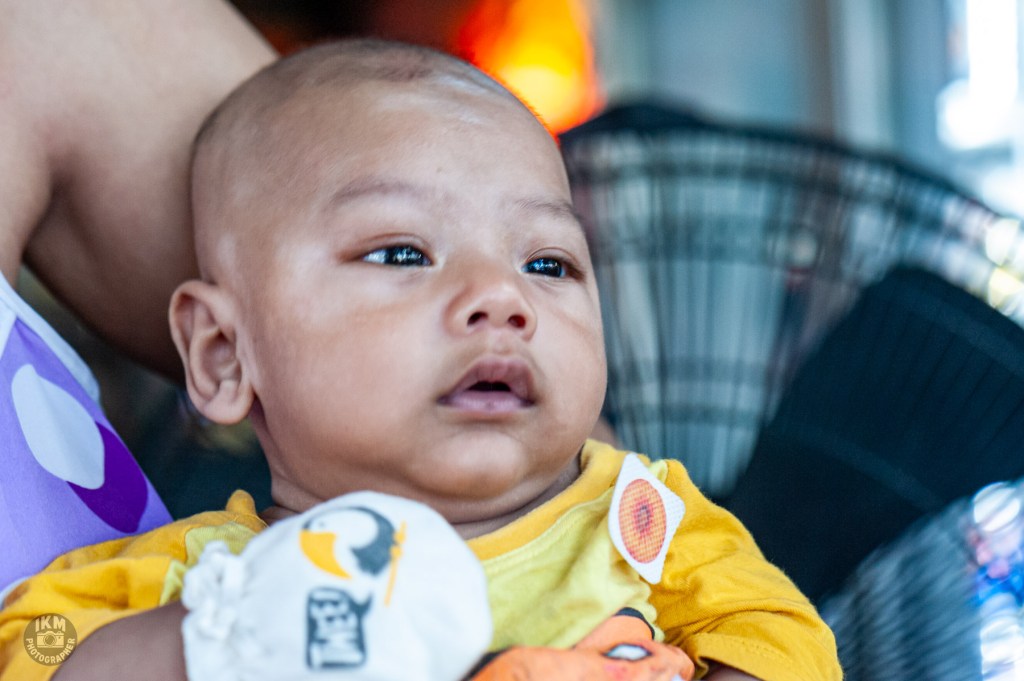 A close-up of a baby with a bald head, wearing a yellow shirt, looking thoughtfully while being held, with a blurred background.