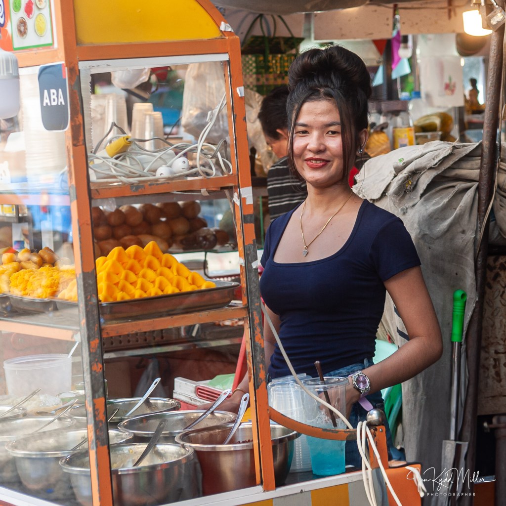 A young woman smiling while standing at a food stall, showcasing colorful desserts and snacks behind her.
