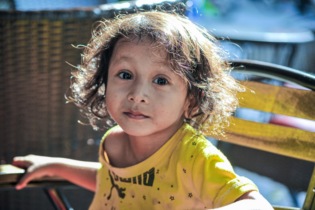 A young child with curly hair sitting at a table, wearing a yellow shirt with black stars, looking directly at the camera.