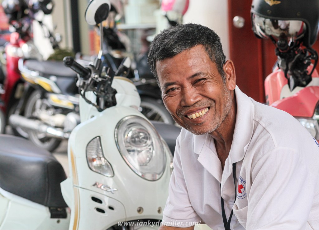A smiling man in a white polo shirt sitting beside a scooter in a parking area, with other motorcycles and helmets visible in the background.